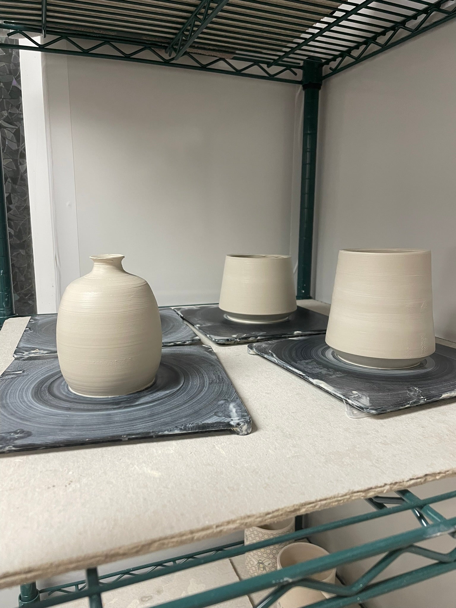 Three damp wheel-thrown clay pieces—a small bottle-shaped vase and two straight-sided cups—resting on square bats on a wire shelving unit in a pottery studio