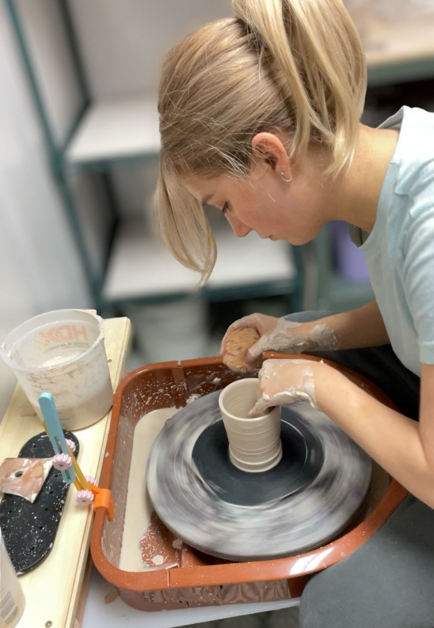 A woman with blonde hair tied back leans over a pottery wheel, shaping a cylindrical clay vessel with her hands; a sponge, brushes, and a water bucket sit nearby on the work surface.